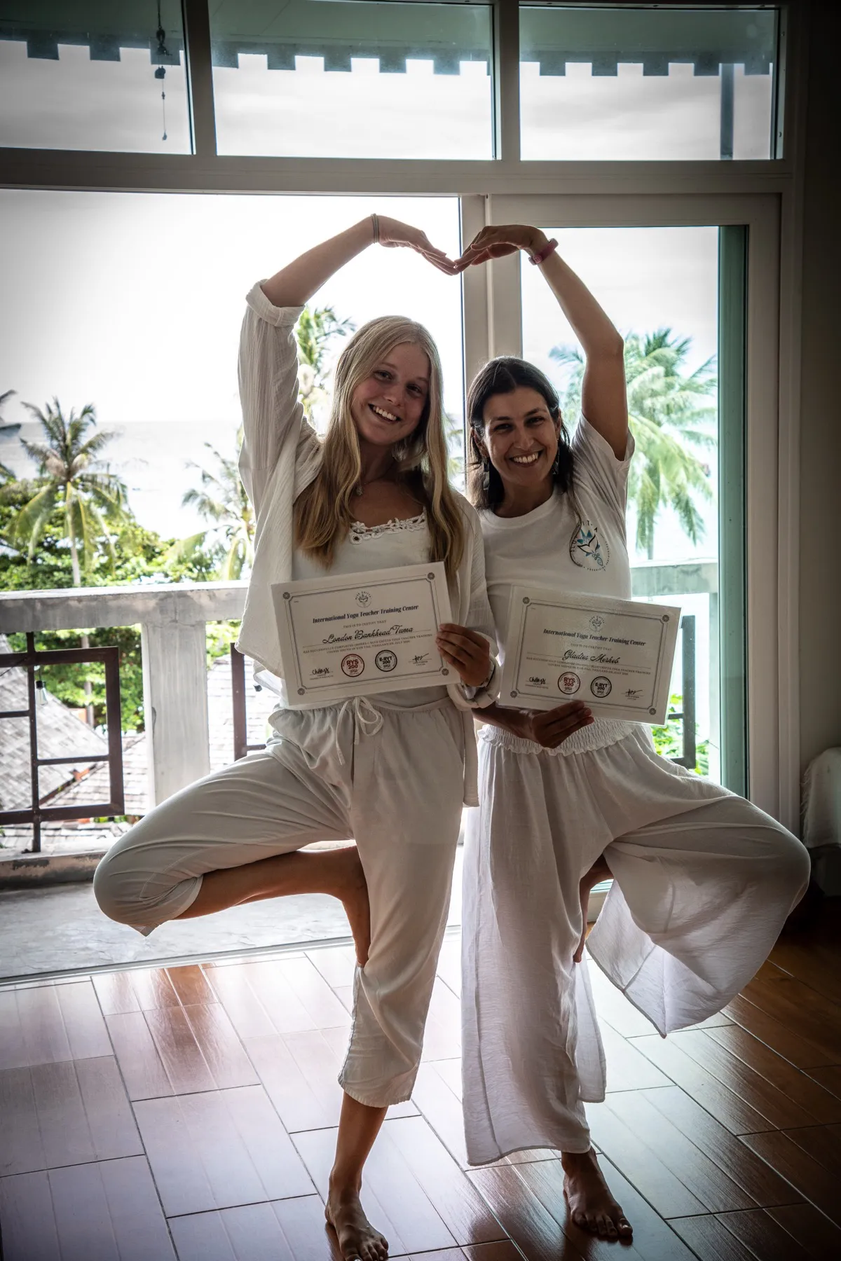 Two graduates in tree pose holding their teacher training certificates